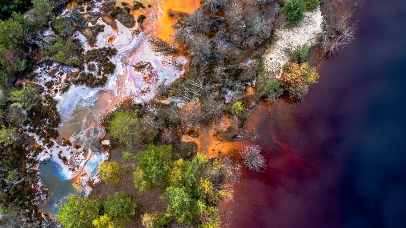 Drohnenaufnahme der Lausitz: rostrot-orangefarbenes Grubenwasser trifft auf dunkelvioletten See, umgeben von Wald